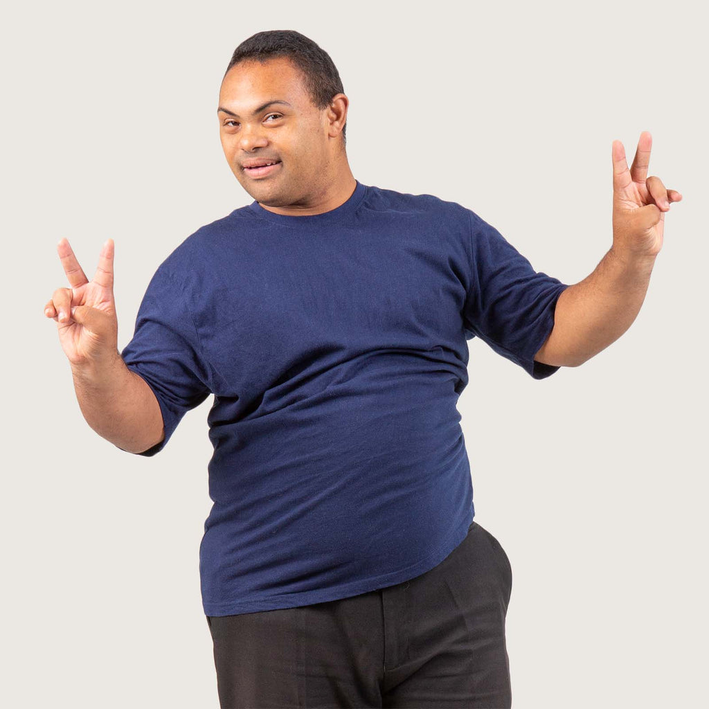 Brad - A young man with Downs syndrome. He is dual heritage and wears a dark blue T-shirt and black trousers. He is holding up two victory signs and smiling.