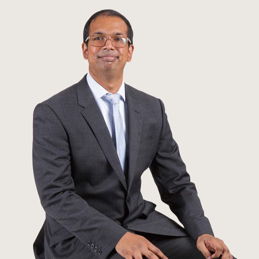 Dinesh - A young British-Indian man with short hair and glasses. He wears a grey suit and tie and is sitting comfortably and looking at the camera.