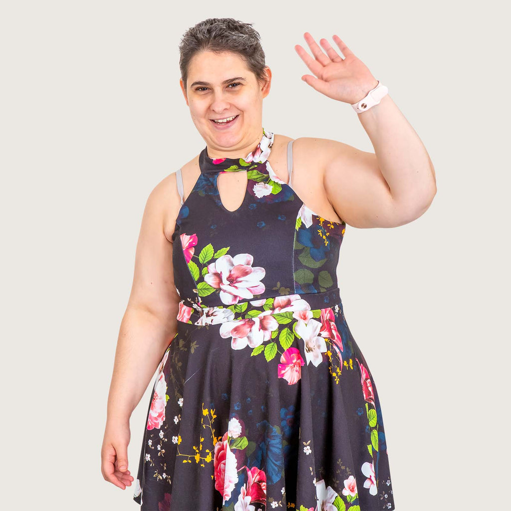 Eva - A young woman with Greek heritage, wearing a bright summer dress with short cropped hair. She is waving at the camera.