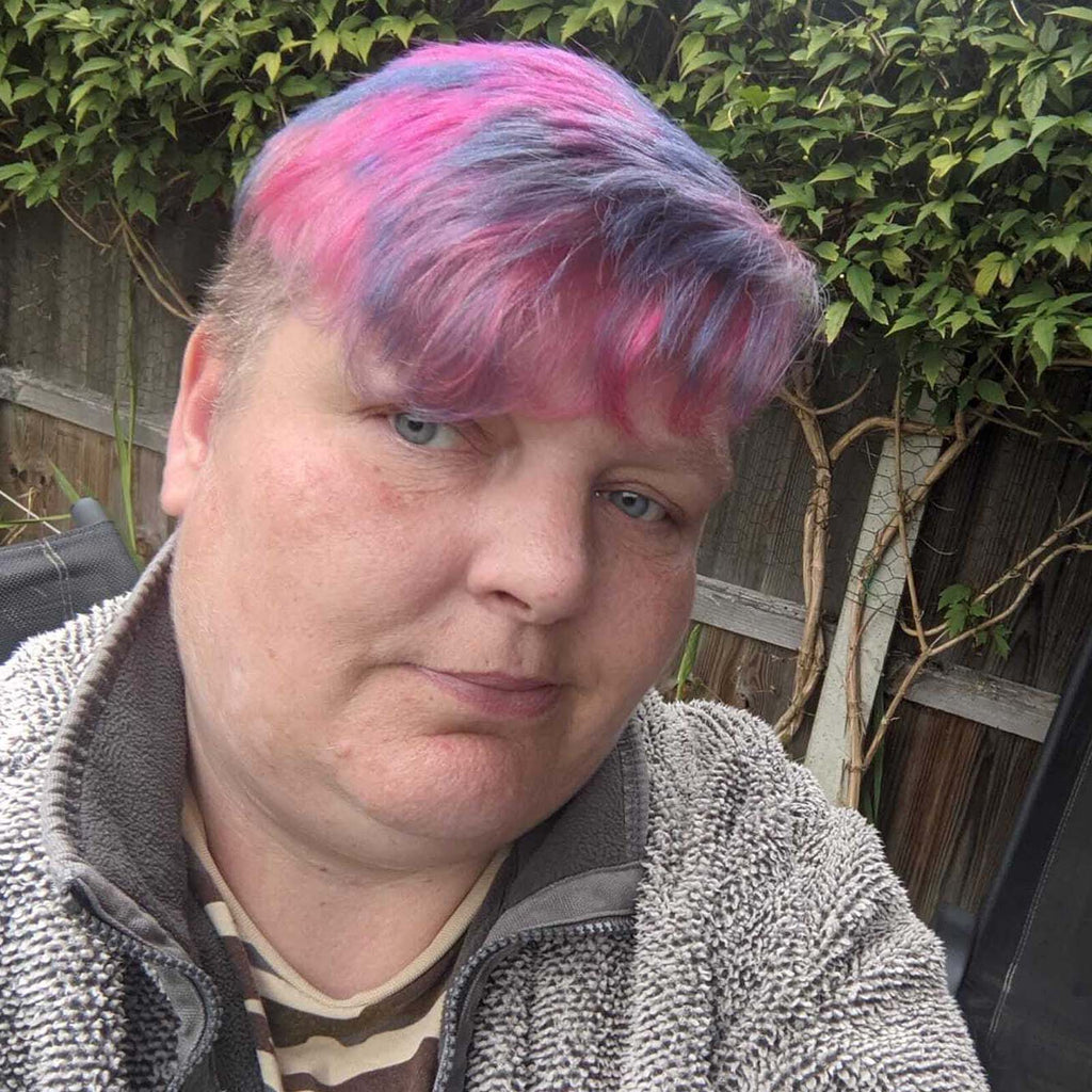 A portrait photo of Hayley - she has short blue and pink hair and wears a grey fleece. She is sat outside in a garden.