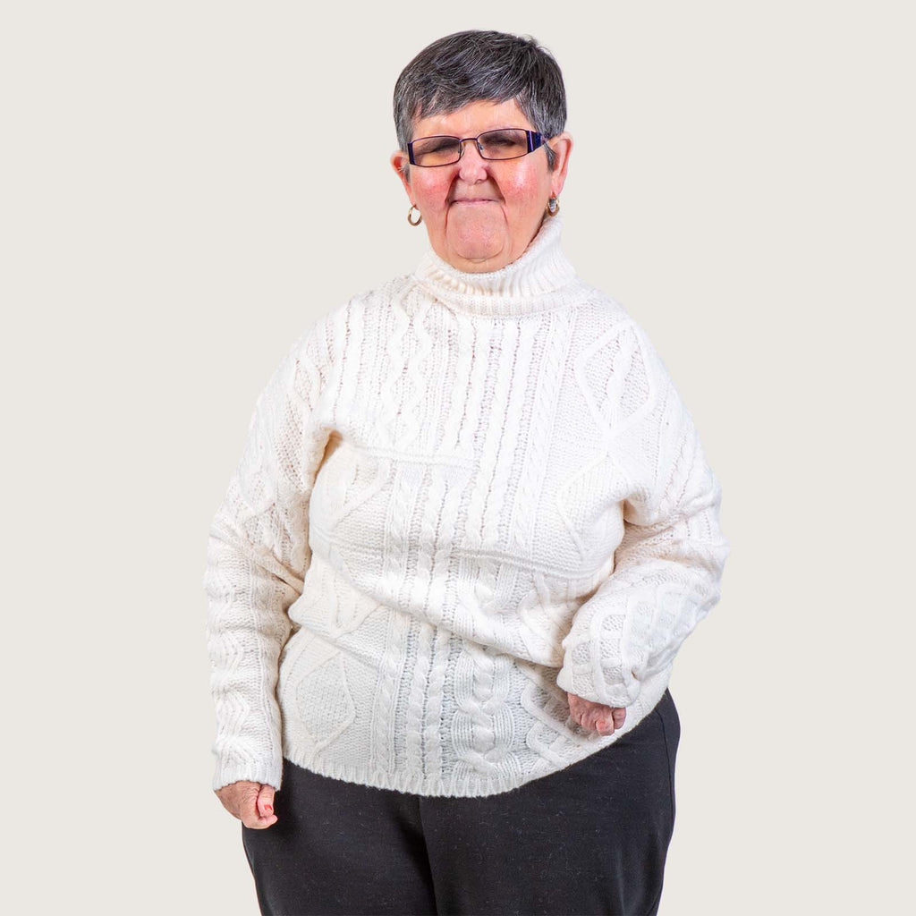 Paula - An older white woman with short grey hair and glasses. She wears a cream jumper and black trousers.