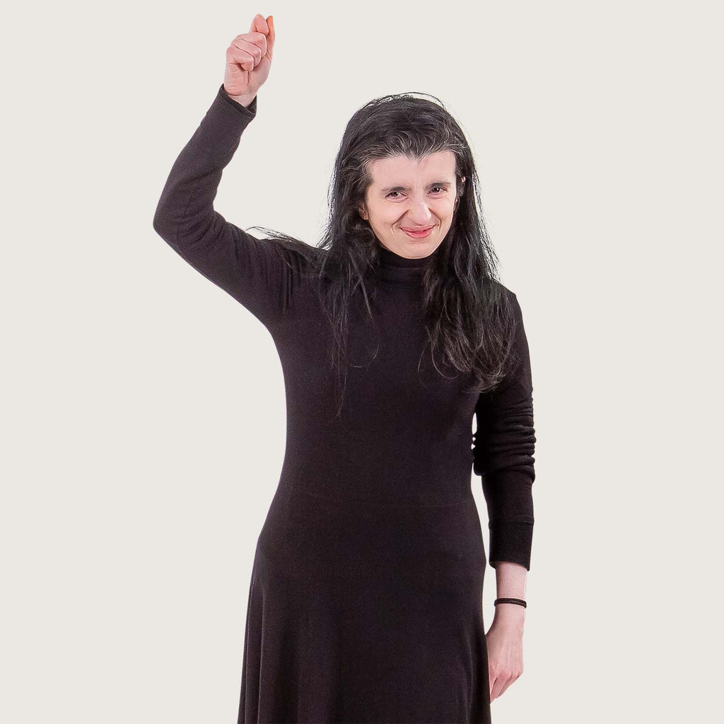 Salma - A young British Bangladeshi woman wears all black. She is smiling and waving at the camera.