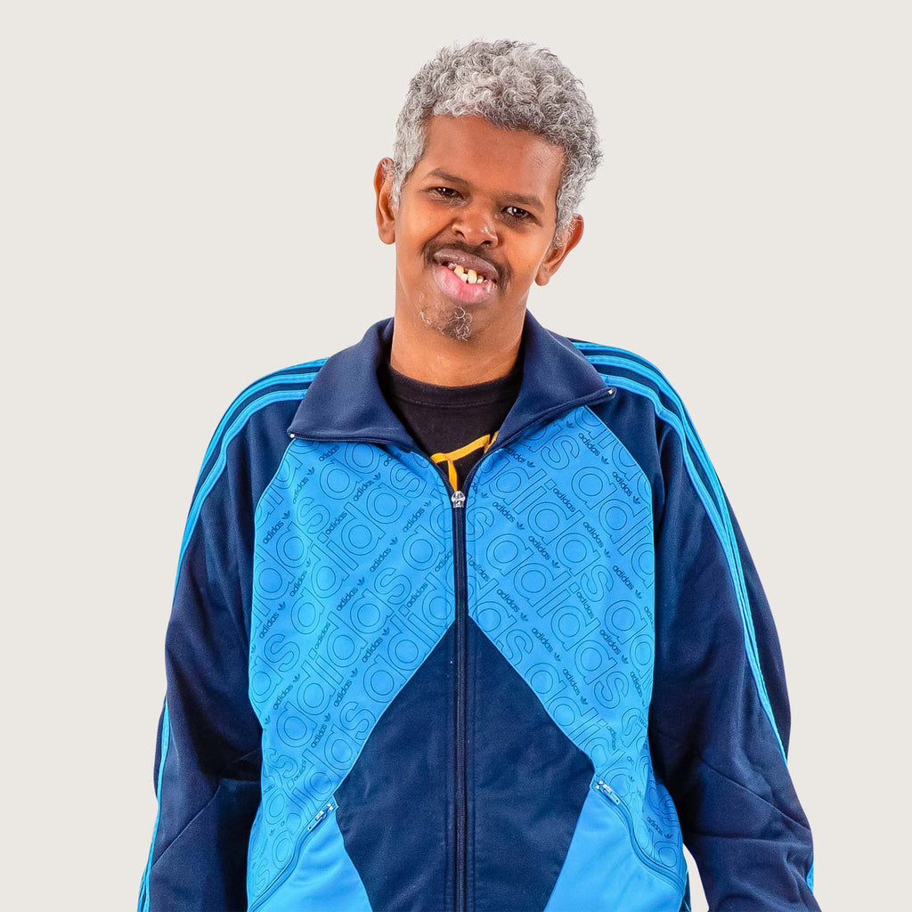 Zahi - A middle aged British-Somali man wears a bright blue tracksuit. He is smiling at the camera.