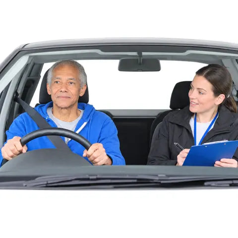 An older man at the wheel of a car, with a driving examiner holding a clipboard in the passenger seat.