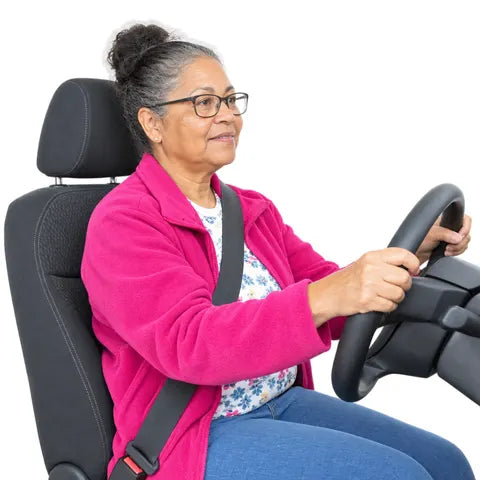 An older woman with glasses and a pink fleece, holding the steering wheel.